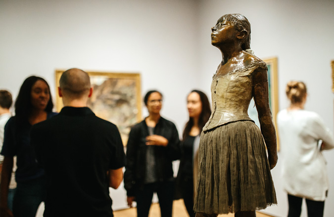 A bronze statue of a young girl is surrounded by visitors gathering for an evening event.