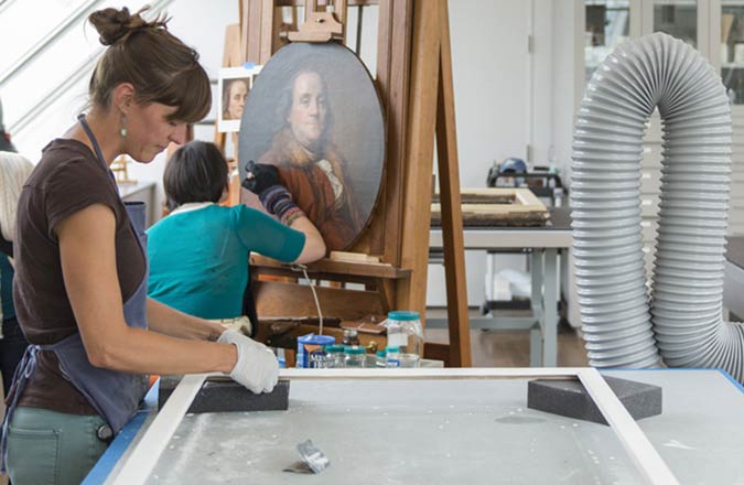 Conservators work on a frame and a portrait of Benjamin Franklin.