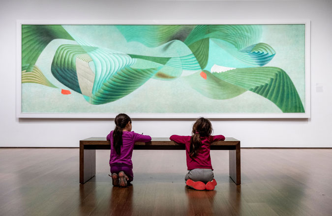 Two children kneel at a gallery bench and draw in front of a large, wide painting with abstract green lines.