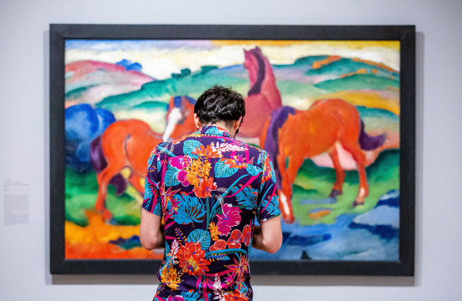 A visitor with a colorful, patterned shirt stands in front of a similarly colorful painting of horses.